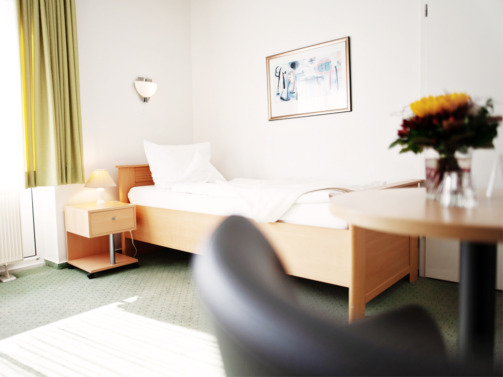 SoVD - helles Einzelzimmer mit grünem Teppich, Einzelbett und Blumenstrauß in einer Vase auf einem Tisch